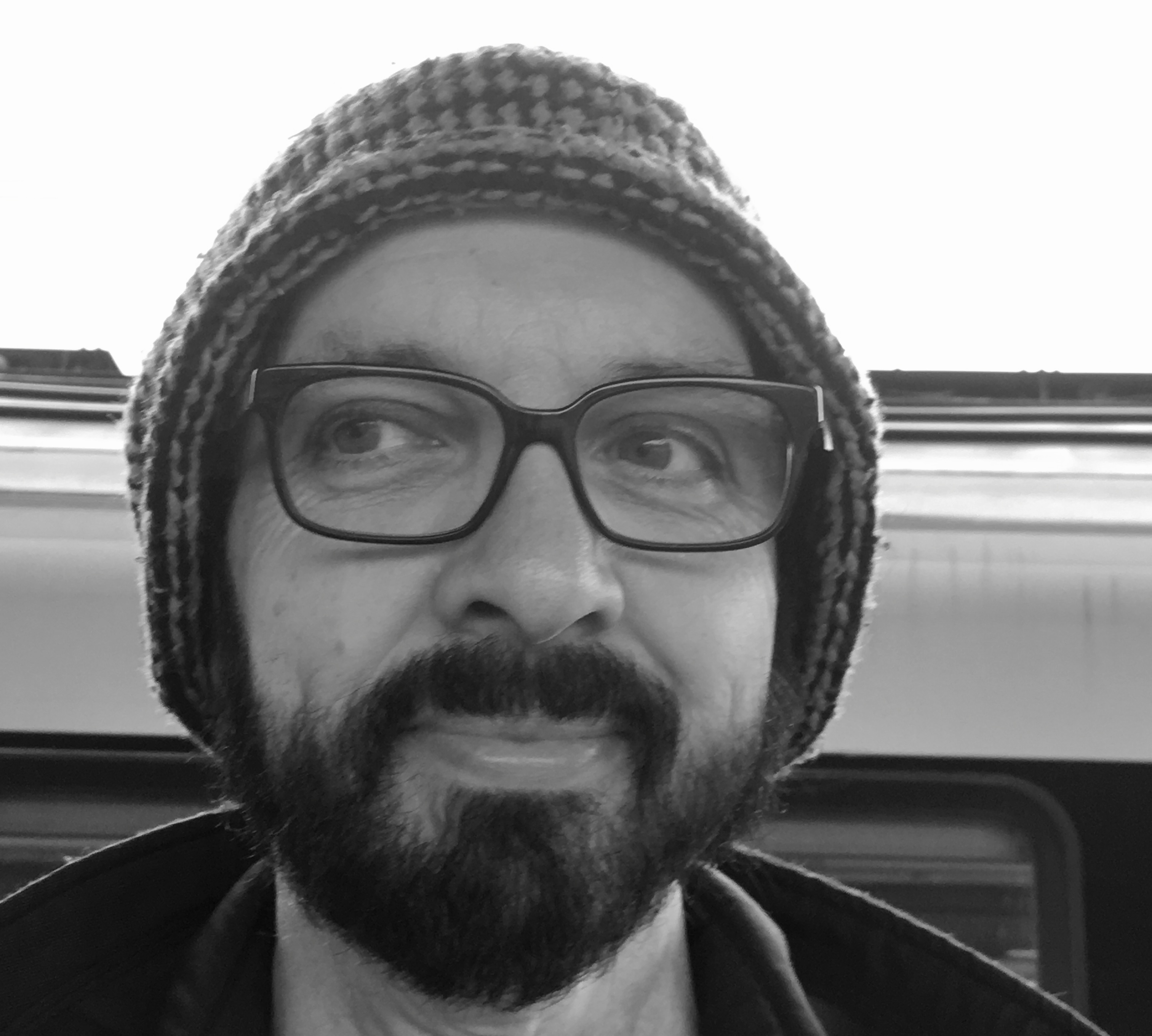 Black and white portrait photo of a man with a beard and glasses smirking and looking to the right. He's also wearing a beanie hat.