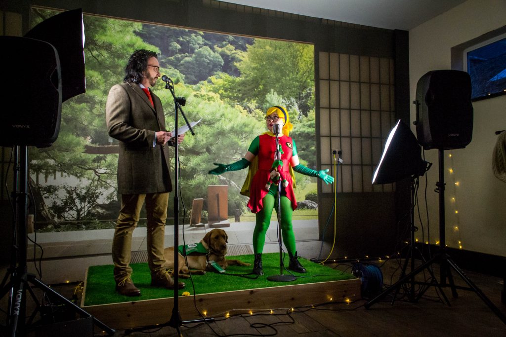 Man and woman stood on a stage. Man is dressed in a suit and the woman is wearing a Robin costume with Yellow wig.