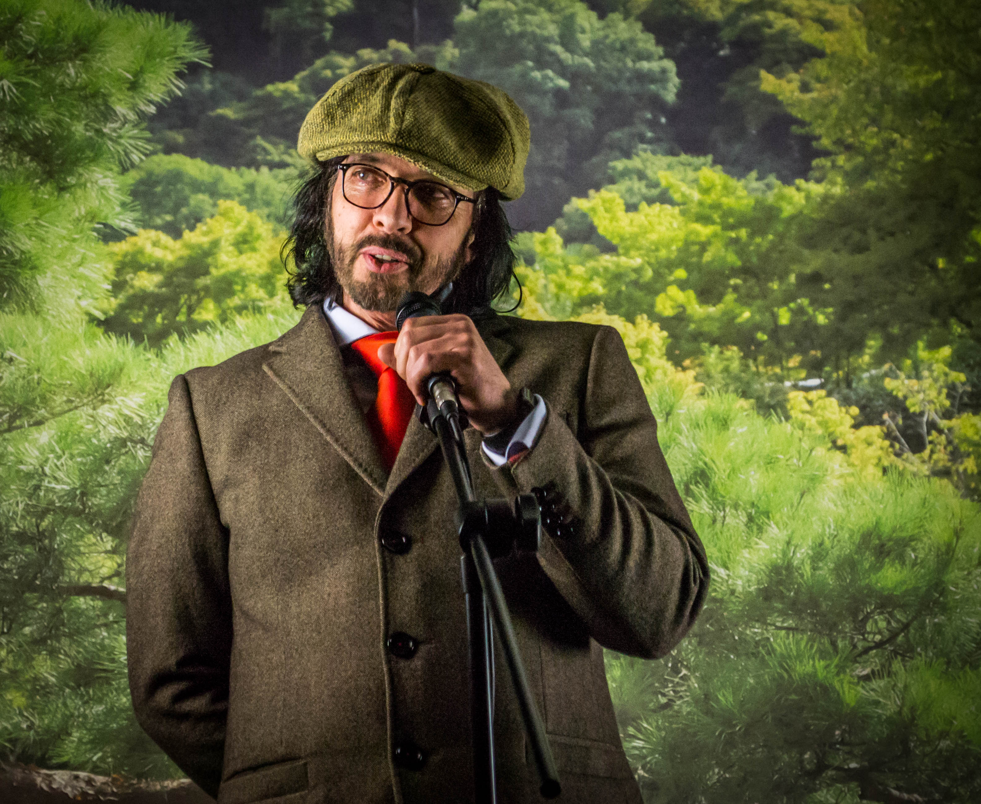 Man in flat cap and smart jacket stood in front of a microphone in front of trees.
